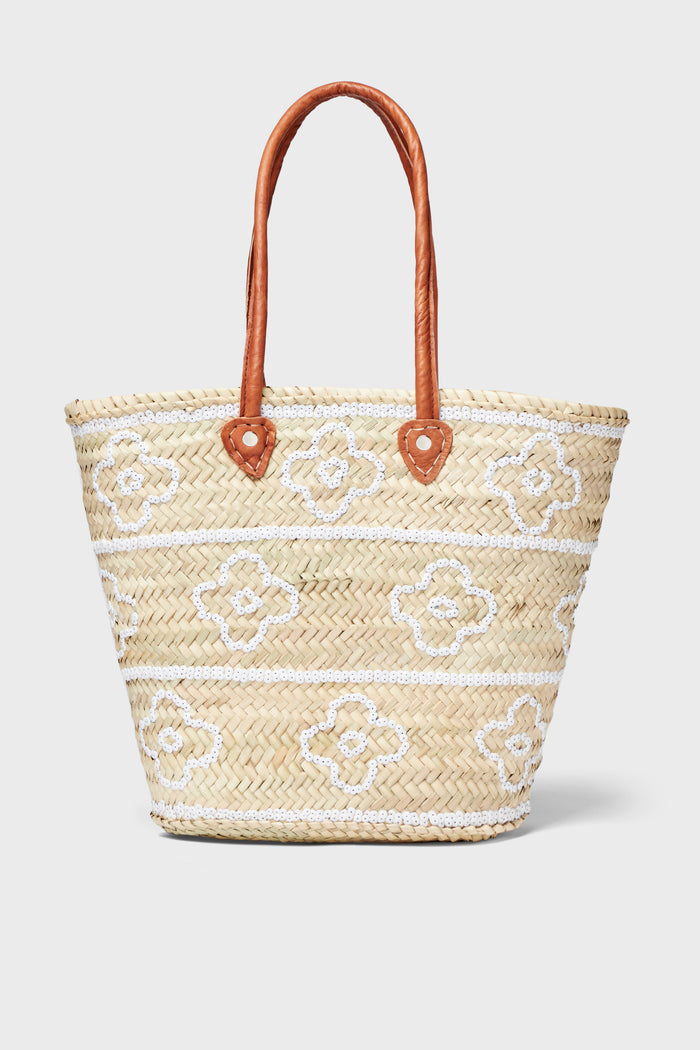 The ASHA x Tuckernuck woven straw bag features decorative white and brown flowers, making it a charming choice for a beach tote.