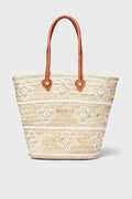 The ASHA x Tuckernuck woven straw bag features decorative white and brown flowers, making it a charming choice for a beach tote.
