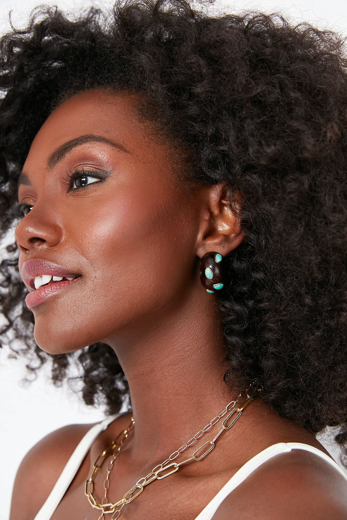 A woman with curly hair wears a white top and Lizzie Fortunato layered gold chain necklaces and handcrafted black earrings with blue accents, looking to the side against a white background.