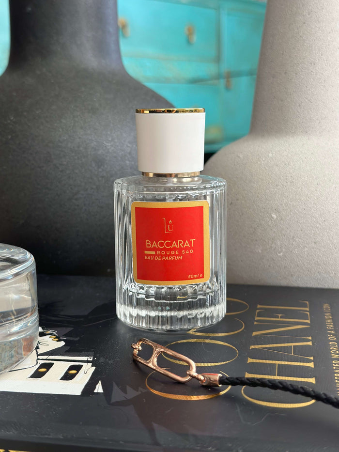Amber & Saffron Eau de Parfum by LU Fragrances — 50ml glass bottle with bold red label and white gold cap, styled on a Coco Chanel coffee table book with a dark ceramic vase and gold chain in the foreground