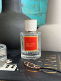 Amber & Saffron Eau de Parfum by LU Fragrances — 50ml glass bottle with bold red label and white gold cap, styled on a Coco Chanel coffee table book with a dark ceramic vase and gold chain in the foreground