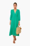 A woman wears the XiRENA Ella Dress in Apple Green—a tiered cotton gauze midi with long sleeves—holding a woven handbag and wearing brown sandals.
