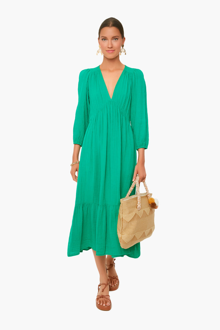 A woman wears the XiRENA Ella Dress in Apple Green—a tiered cotton gauze midi with long sleeves—holding a woven handbag and wearing brown sandals.