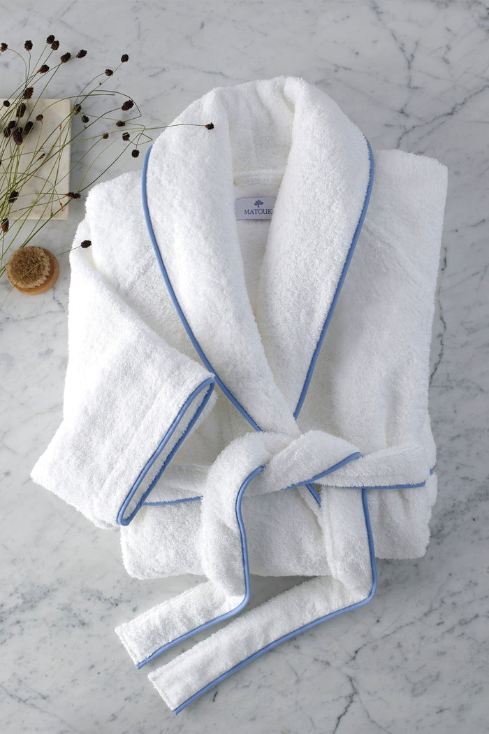 A Matouk white cotton terry robe with light blue trim is neatly folded on marble, paired with a brush and dried plant stems, evoking the luxurious comfort of high-end resort linens.
