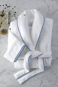 A Matouk white cotton terry robe with light blue trim is neatly folded on marble, paired with a brush and dried plant stems, evoking the luxurious comfort of high-end resort linens.