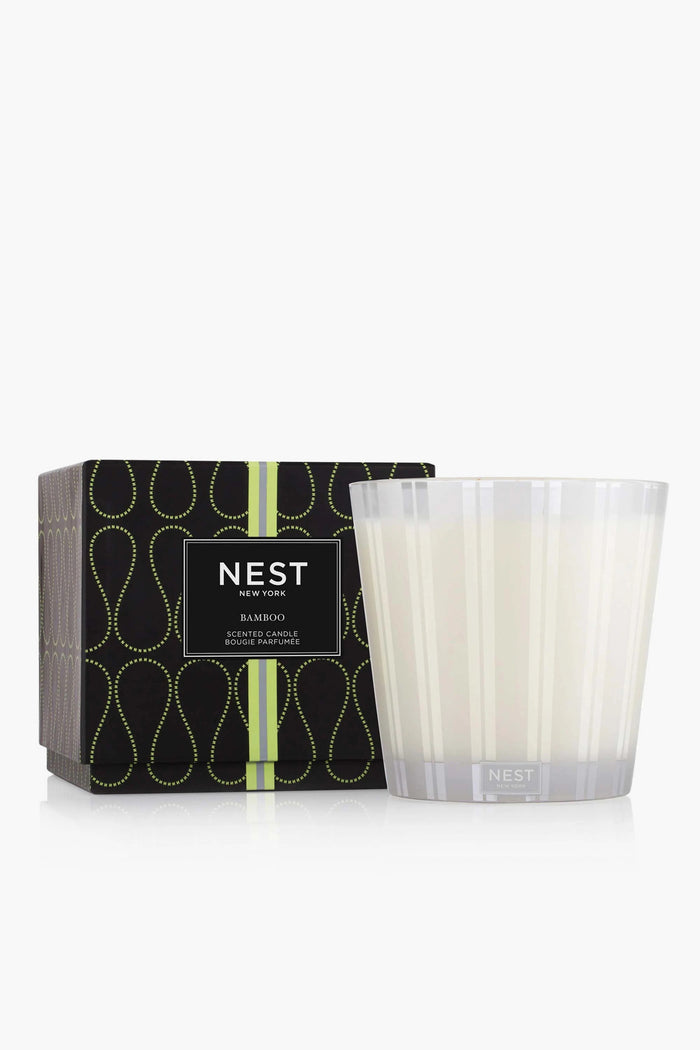 A NEST New York luxury candle with a bamboo scent sits in a clear striped glass holder next to its black decorative box with green accents.