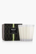 A NEST New York luxury candle with a bamboo scent sits in a clear striped glass holder next to its black decorative box with green accents.