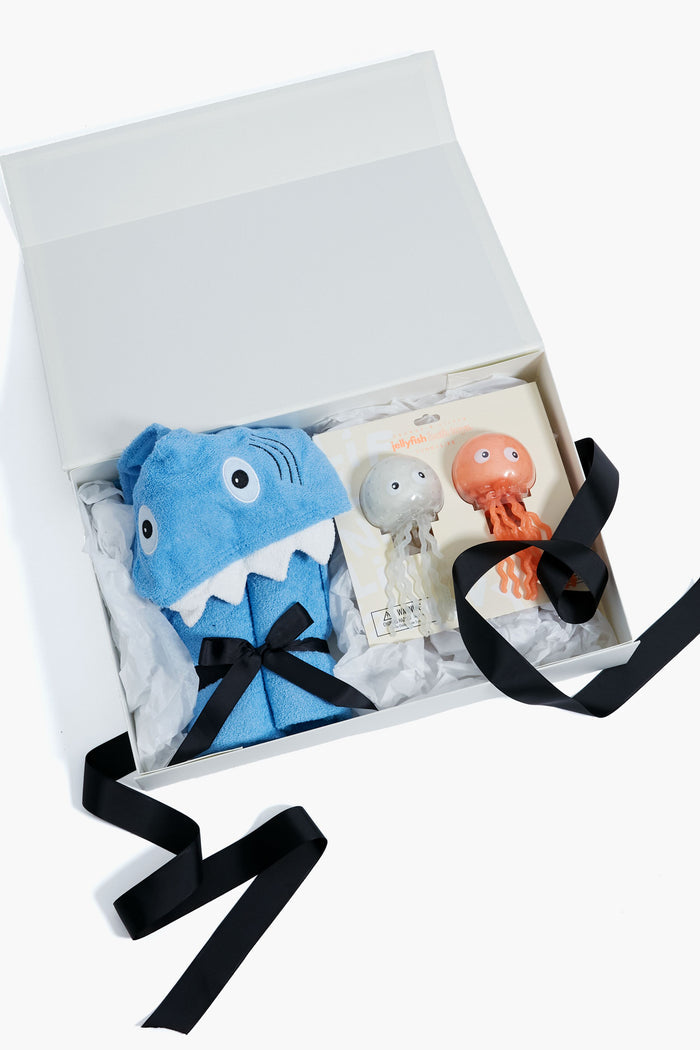 Tuckernuck Tots gift box with a blue shark-themed hooded towel and two bath squirters, finished with black ribbon—perfect for toddler gifts.