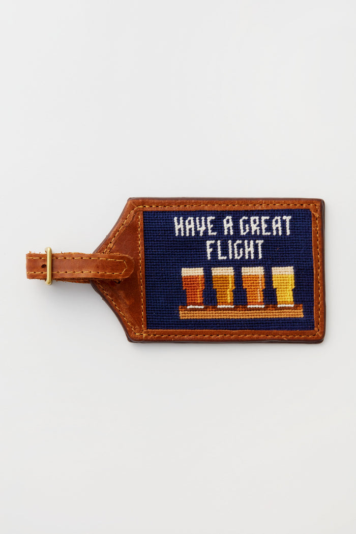 The Smathers and Branson brown leather luggage tag features hand-stitched needlepoint with "Have a Great Flight" above four beer glasses.