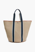 A Rue de Verneuil beige luxury tote bag with dark blue and white striped handles and brown trimmed edges stands upright.