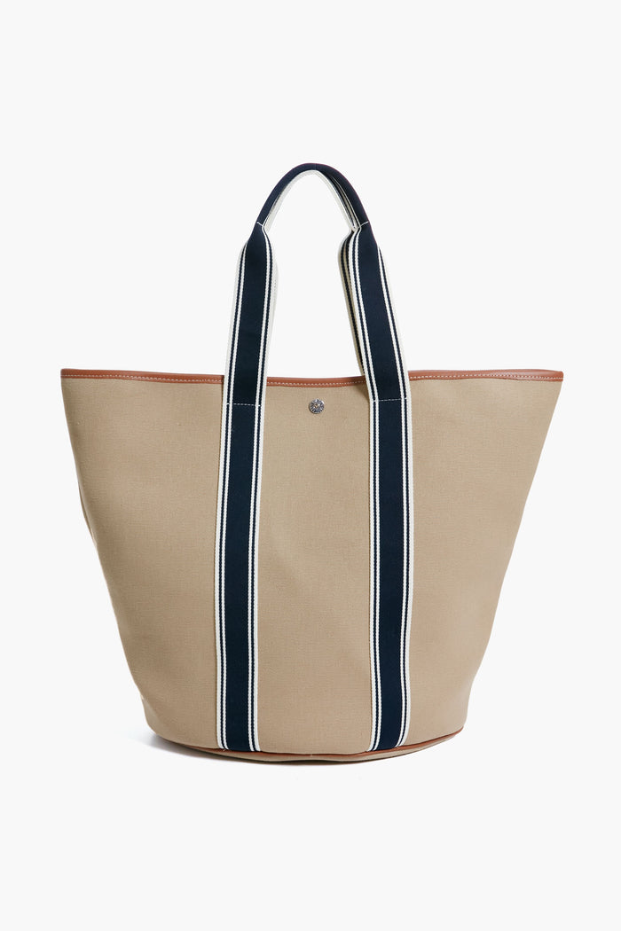 A Rue de Verneuil beige luxury tote bag with dark blue and white striped handles and brown trimmed edges stands upright.