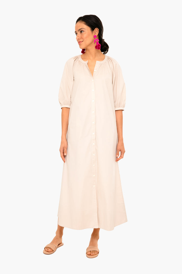 A woman wears the STAUD Vincent Dress—a light beige, button-down caftan with three-quarter sleeves—styled with pink statement earrings and flat sandals for an effortless look.
