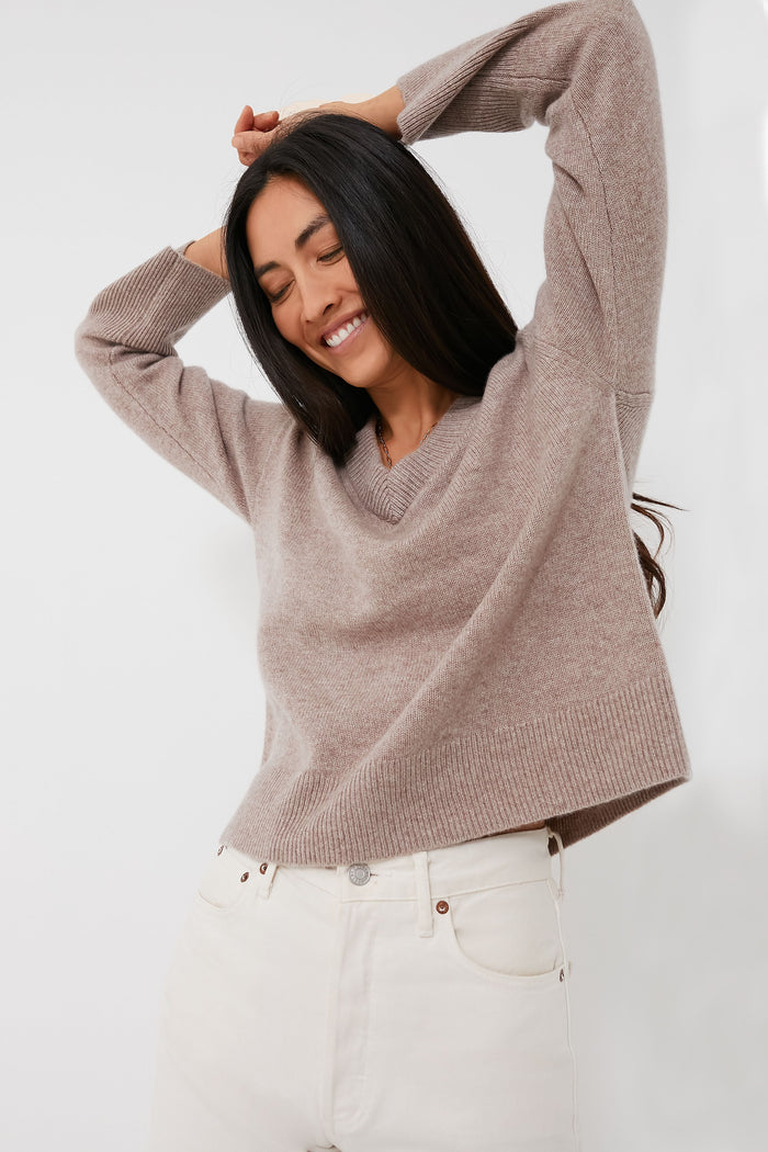 A woman in an Alex Mill beige V-neck knit sweater and white jeans poses with arms raised, smiling with her eyes closed.