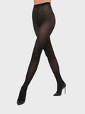 A person poses in a minimalist setting wearing geometric pattern black tights from Wolford and black high-heeled shoes.