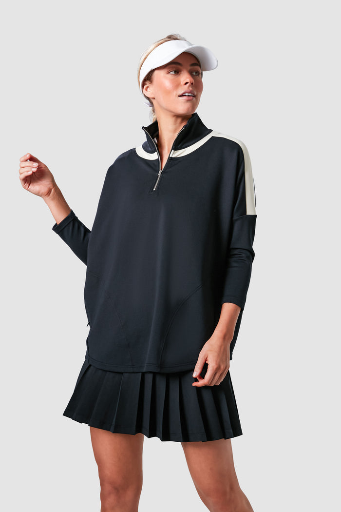 A woman in Tnuck Sport's black half-zip pullover with white accents, pleated black skirt, and white visor showcases cooling fabric for an athletic tennis dress style.