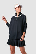 A woman in Tnuck Sport's black half-zip pullover with white accents, pleated black skirt, and white visor showcases cooling fabric for an athletic tennis dress style.