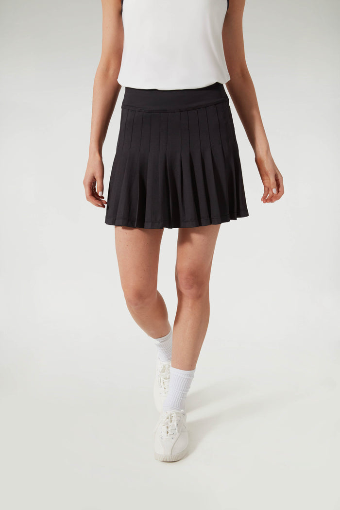 A person in Tnuck Sport’s white sleeveless top, pleated tennis skirt, white socks, and white sneakers stands confidently.