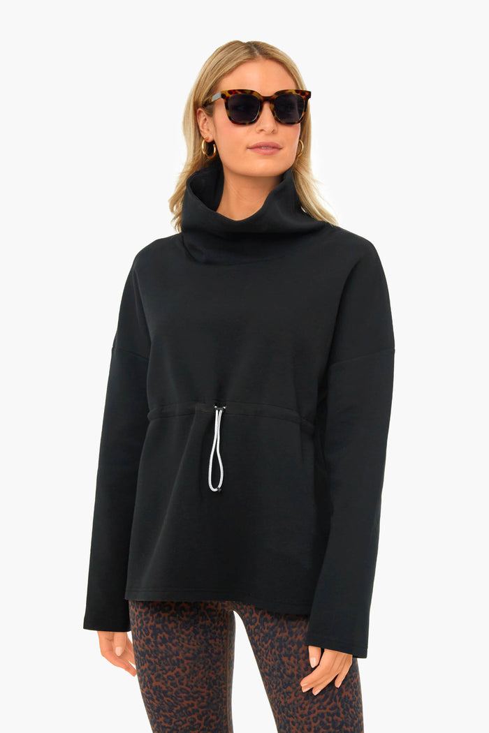 Wearing Varley, a woman in sunglasses showcases a black long-sleeve top with front drawstring, funnel collar, and patterned leggings, embodying performance-fashion with effortless style.