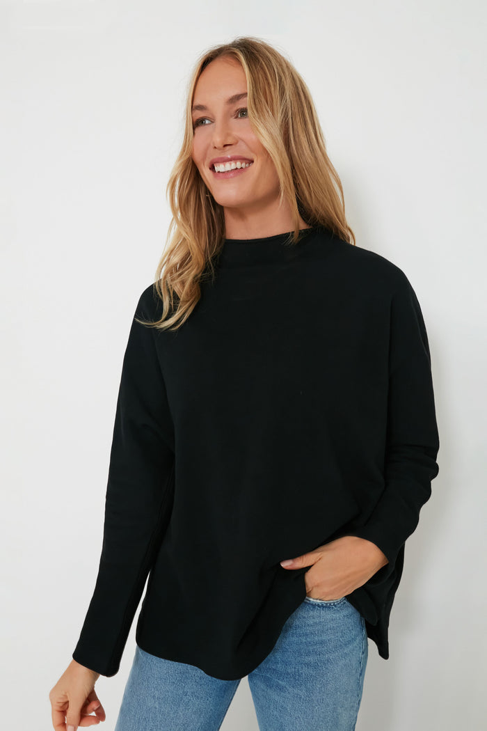 A person with long blonde hair is smiling, wearing a Pomander Place black long-sleeve terry-style pullover and blue jeans.
