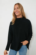 A person with long blonde hair is smiling, wearing a Pomander Place black long-sleeve terry-style pullover and blue jeans.