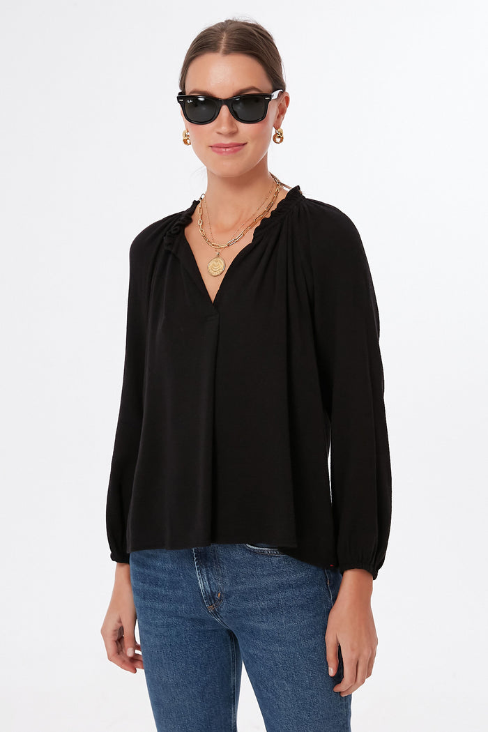 A person wearing sunglasses, gold jewelry, blue jeans, and a XiRENA Black Camille Top with a ruffled collar stands confidently.