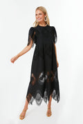 A woman wears Tuckernuck's Catalina Dress—a black midi dress with lace cutouts, geometric patterns, short sleeves—paired with gold sandals and large hoop earrings.