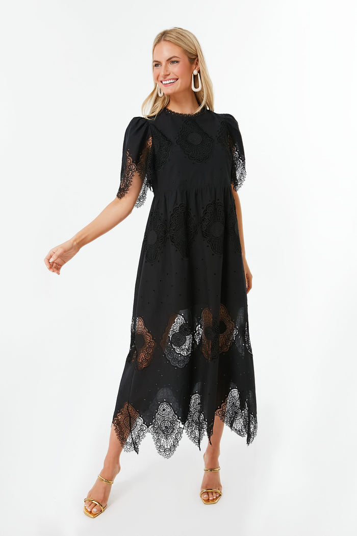 A woman wears Tuckernuck's Catalina Dress—a black midi dress with lace cutouts, geometric patterns, short sleeves—paired with gold sandals and large hoop earrings.