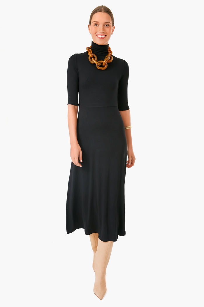 A woman wears a Vince black turtleneck midi dress with short sleeves, paired with a chunky brown necklace, gold bracelet, and beige knee-high boots.