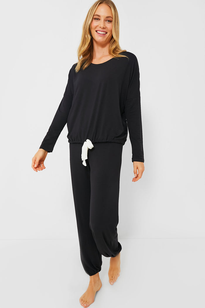 A woman wearing a long-sleeve black modal jersey pajama set with a white tie waist from Eberjey stands barefoot, smiling at the camera.