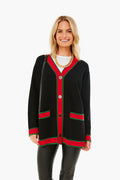 A woman stands and smiles in a Tuckernuck oversized black cardigan with red and green trim, gold crest buttons, paired with a gold chain necklace, white shirt, and black pants. This wool piece adds chic style to any outfit.