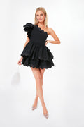 A woman wears a CeliaB black lava dress featuring an asymmetrical neckline and ruffled, layered skirt, paired with a white beaded handbag and clear embellished heels.