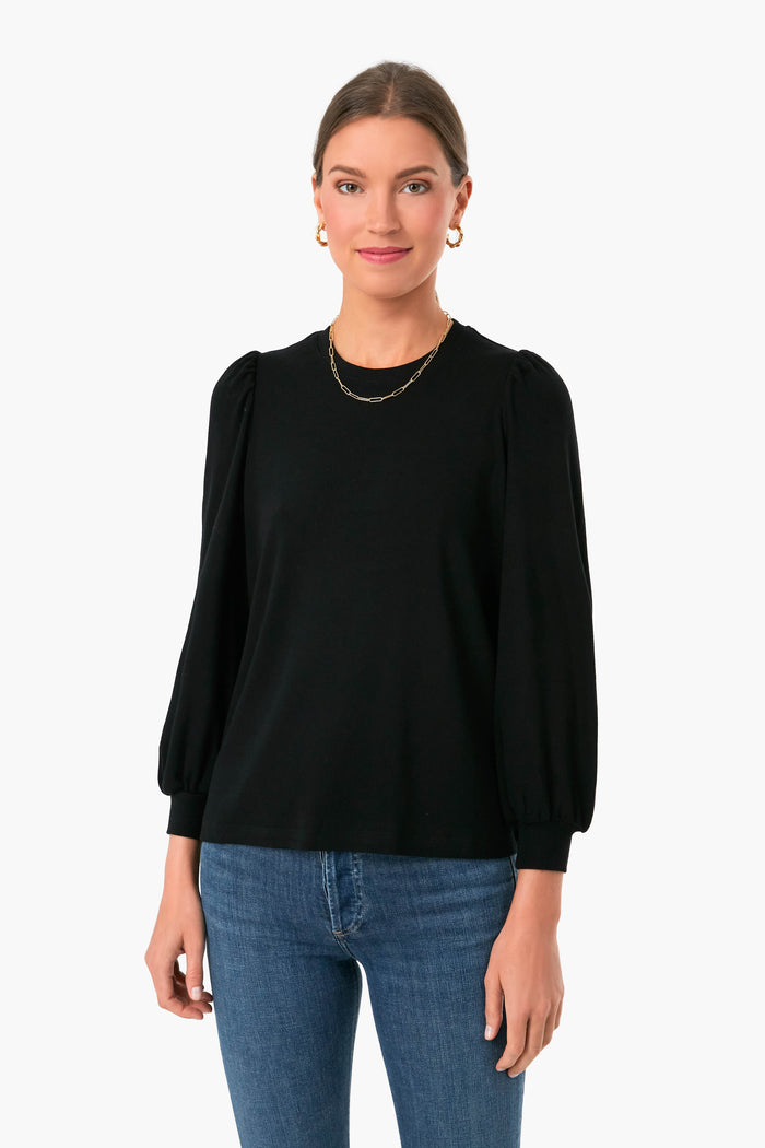 A woman faces forward wearing a Marine Layer black long sleeve rib puff sleeve top and blue jeans, her hair pulled back and accessorized with gold earrings and a necklace, highlighting her preference for sustainably-made fashion.