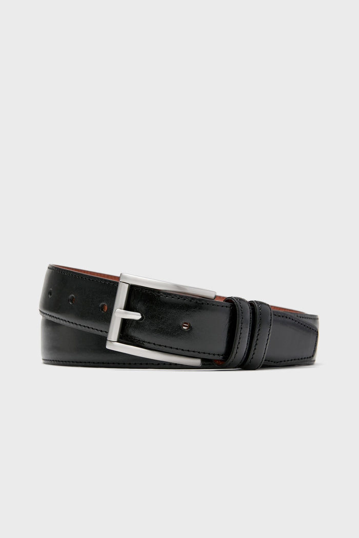 A black leather belt from Trafalgar, featuring a silver metal buckle, is coiled to showcase men's luxury accessories made from full grain leather.