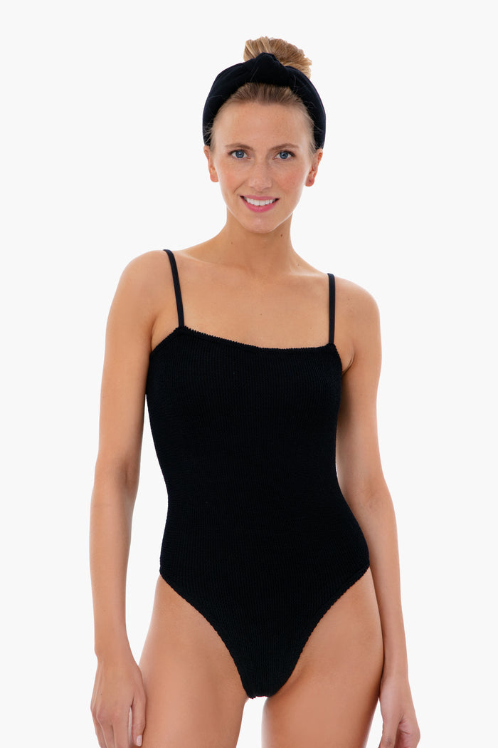 A woman wearing a black Hunza G high-cut one-piece swimsuit with a matching headband smiles at the camera.