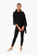 A woman wears a black 100% acrylic layering poncho from Pomander Place, styled with black leggings, gold flats, and a brown clutch.