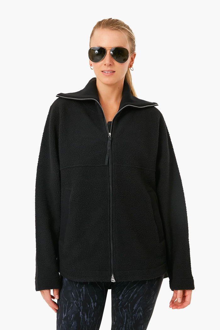 A person in a Varley oversized zip fleece jacket, black patterned leggings, and sunglasses stands facing the camera.