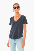 A person stands confidently wearing sunglasses, gold jewelry, a Z SUPPLY Black V Neck Tee made from organic cotton, and light blue jeans.