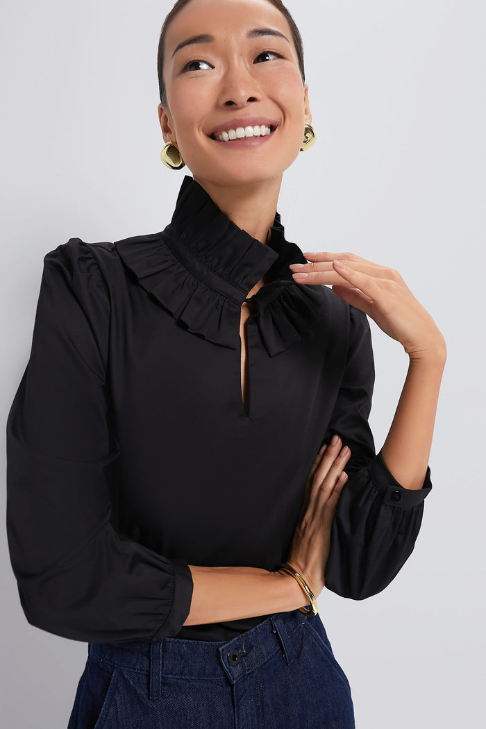A woman in a Tuckernuck black ruffle neck blouse, paired with gold hoop earrings and denim jeans, smiles warmly with one hand resting near her collar.