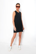 A woman in a sleeveless black Z SUPPLY Sloane dress, white sneakers, sunglasses, and hoop earrings stands confidently, showcasing effortless style.