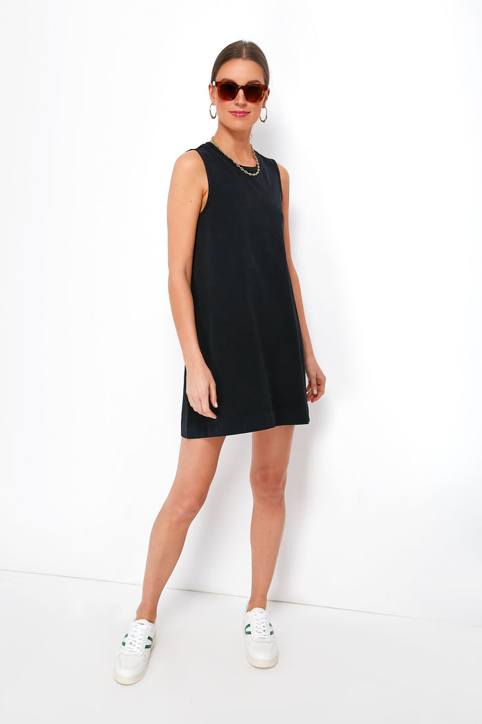 A woman in a sleeveless black Z SUPPLY Sloane dress, white sneakers, sunglasses, and hoop earrings stands confidently, showcasing effortless style.