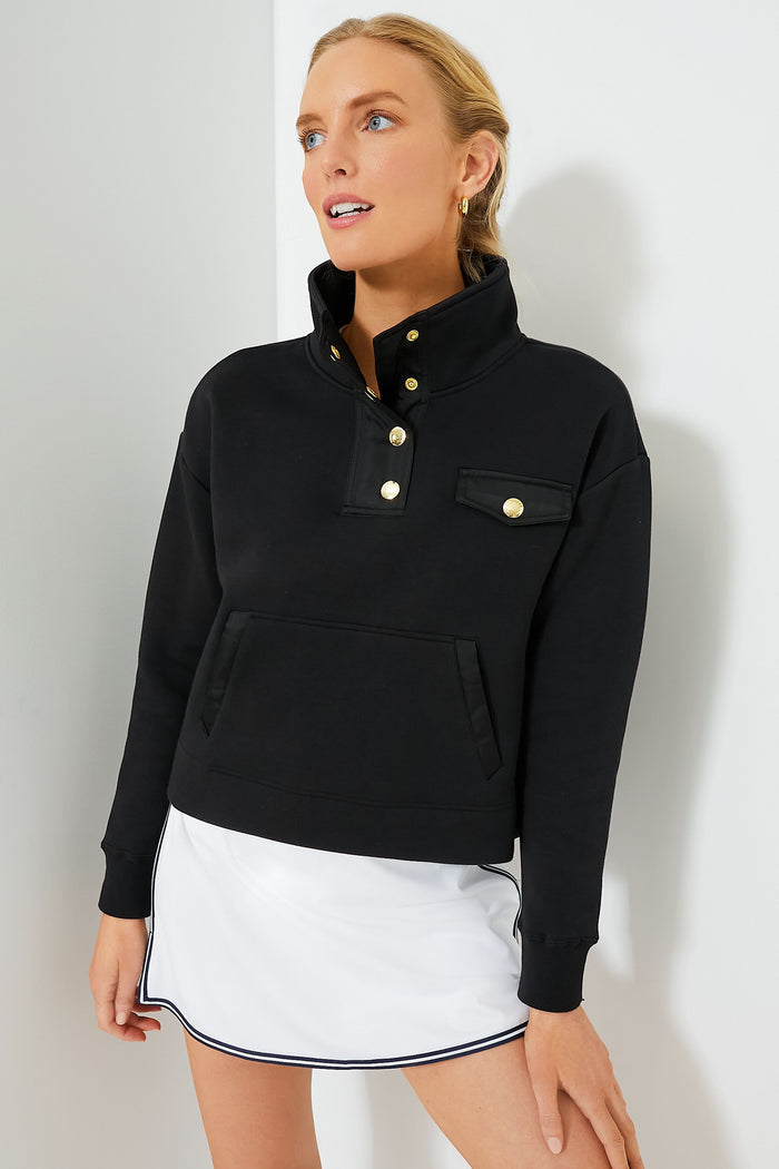 A woman models a Tnuck Sport black snap neck pullover with gold buttons, paired with a white skirt with black trim. Ideal for those seeking stylish ladies’ pullovers.