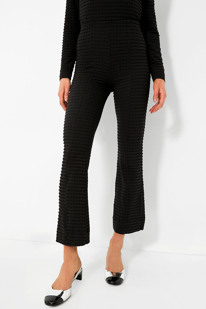 A person wears black textured GANNI pants with a matching long-sleeve top, styled with black and white low-heeled shoes.