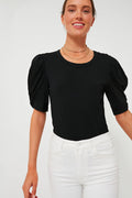 A woman in a Pomander Place Black Vesty Puff Sleeve Crewneck, layered gold necklaces, and high-waisted white jeans stands confidently, smiling.