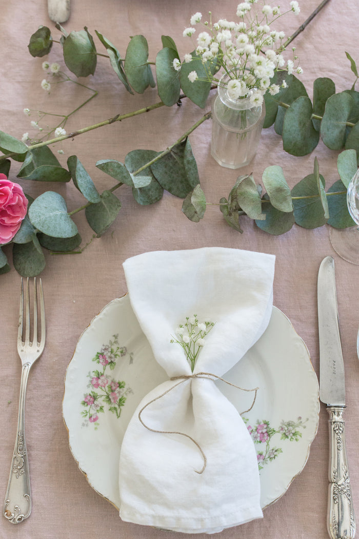 A table is set with a floral plate, fork, and knife. Madame de la Maison Blanc Linen Napkins, tied with twine and small flowers, rest on the plate. Eucalyptus and blooms add charm to these sustainable table linens.