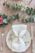 A table is set with a floral plate, fork, and knife. Madame de la Maison Blanc Linen Napkins, tied with twine and small flowers, rest on the plate. Eucalyptus and blooms add charm to these sustainable table linens.