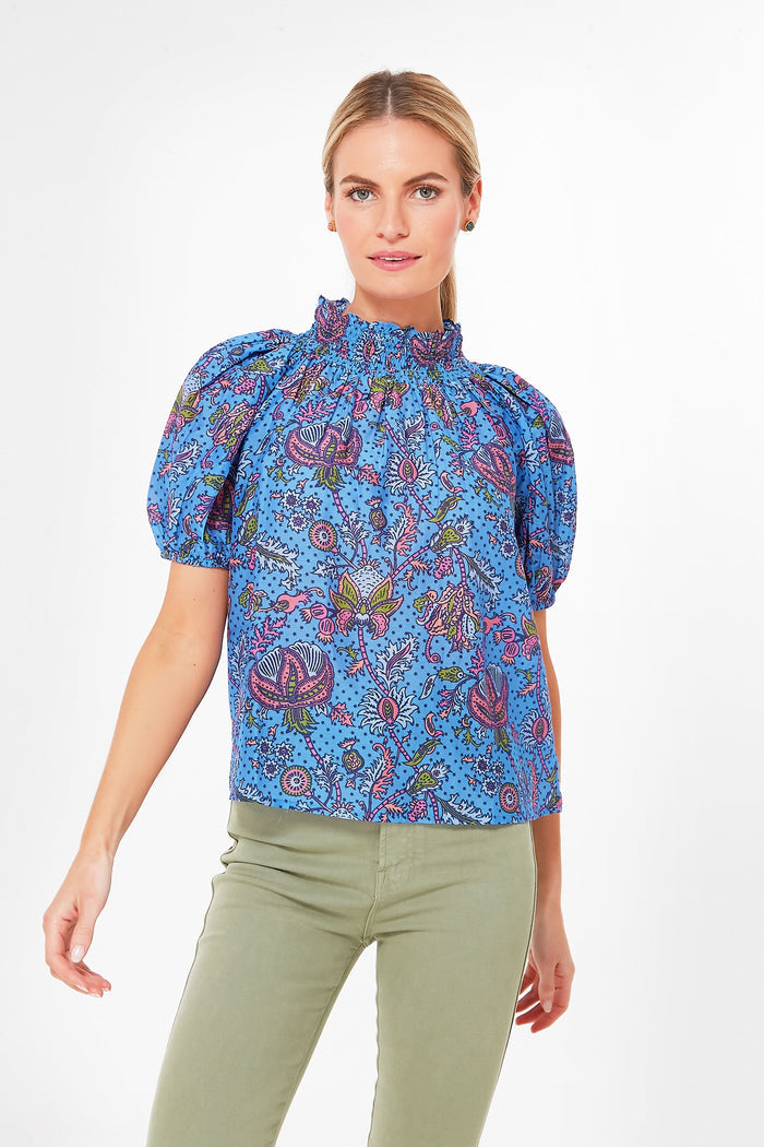 A woman wears a blue paisley printed top with a ruffled neckline and light green pants by Roller Rabbit.