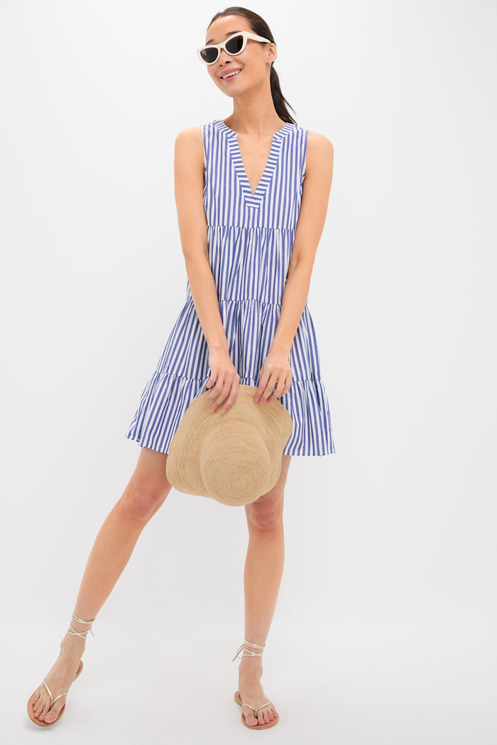 Woman wearing a sleeveless blue and white cotton poplin Pomander Place dress with stripes, white sunglasses, and sandals, holding a straw hat.