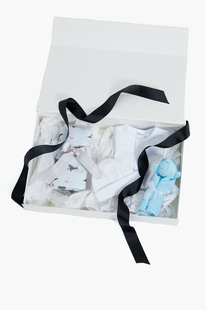 A white Tuckernuck Tots gift box with a black ribbon features folded baby clothes, a patterned item tied with ribbon, and a blue plush toy—an adorable newborn or baby boy gift bundle presented on white tissue paper.