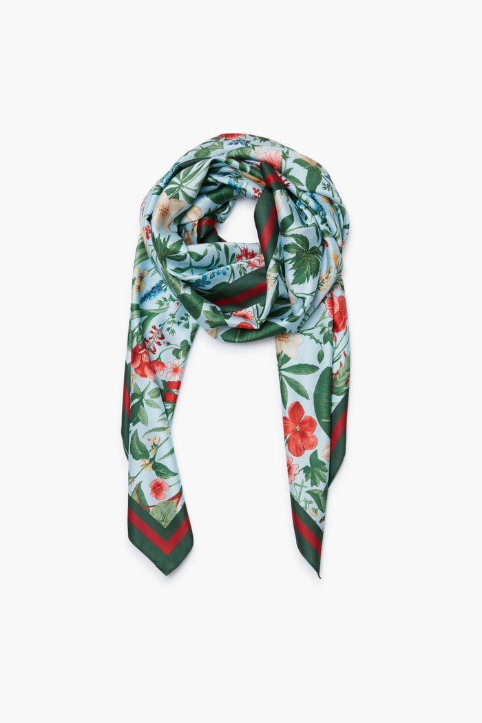 A Tuckernuck Accessories silk Blue Blooming Botanical Scarf, featuring a vibrant floral print in red, green, and blue hues, is styled in a loose knot—an elegant choice for a luxe accessory or those desiring a custom look.