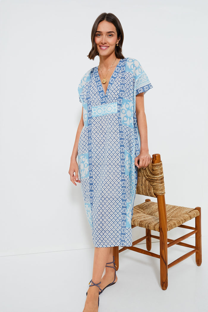 A woman in a light blue and white hand block printed caftan from Love Tomas by Emerson Fry stands beside a woven wooden chair, embodying effortless style.